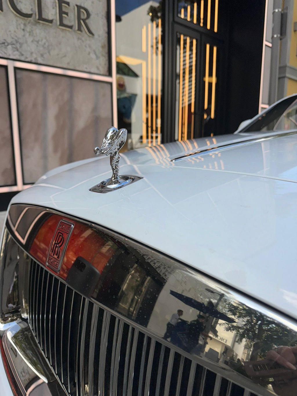 Close-up of a car hood with a prominent emblem, likely a Rolls-Royce, in an urban setting.