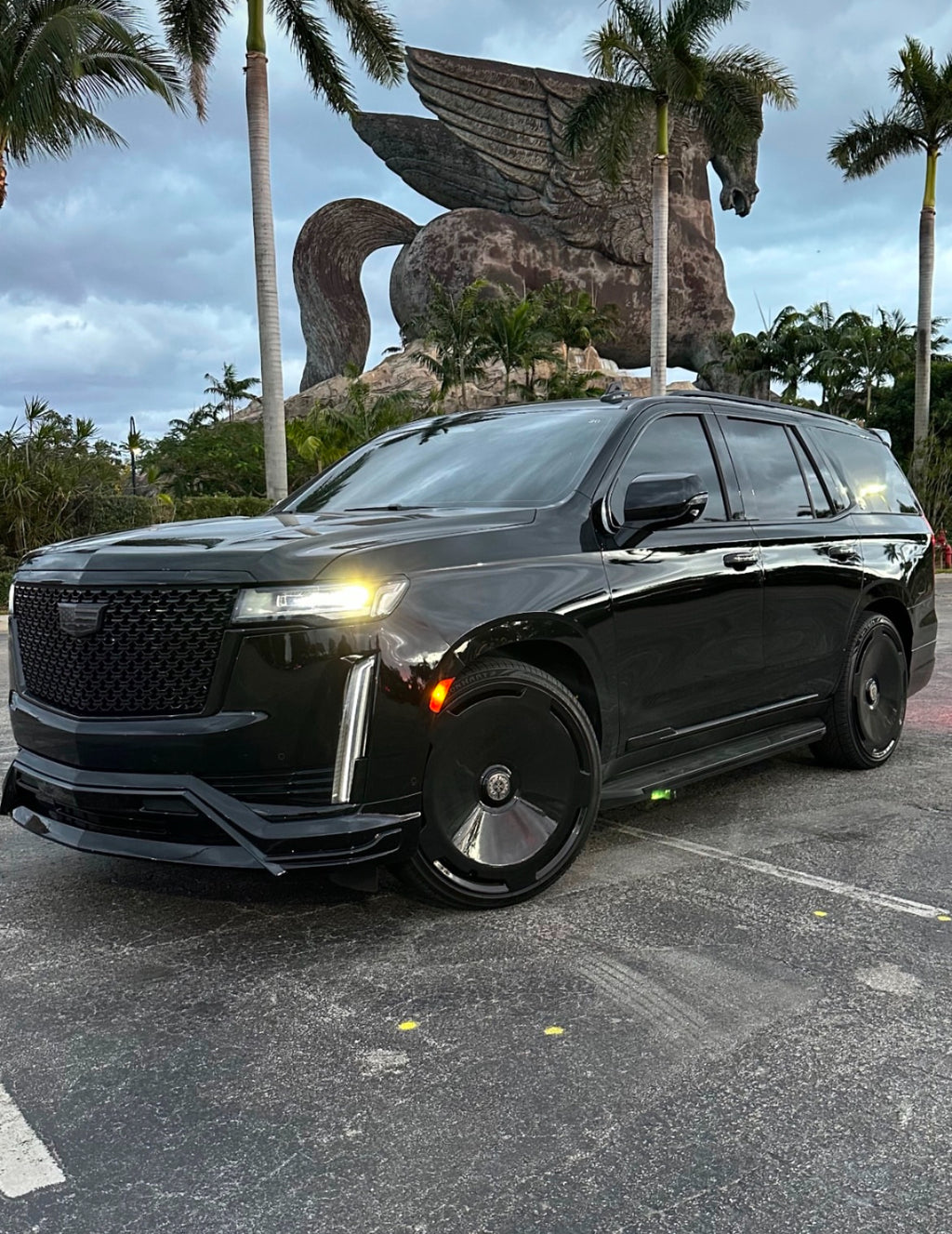 Black Cadillac SUV parked in front of a large statue of a winged horse with palm trees in the background.