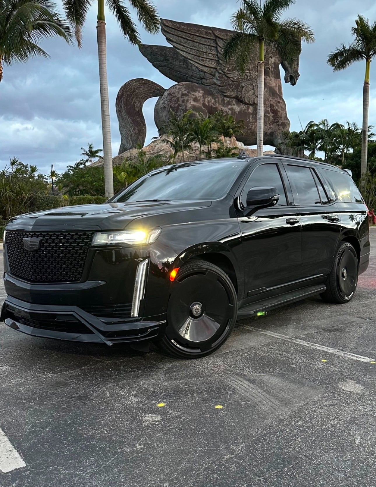 Black Cadillac SUV parked in front of a large statue of a winged horse with palm trees in the background.