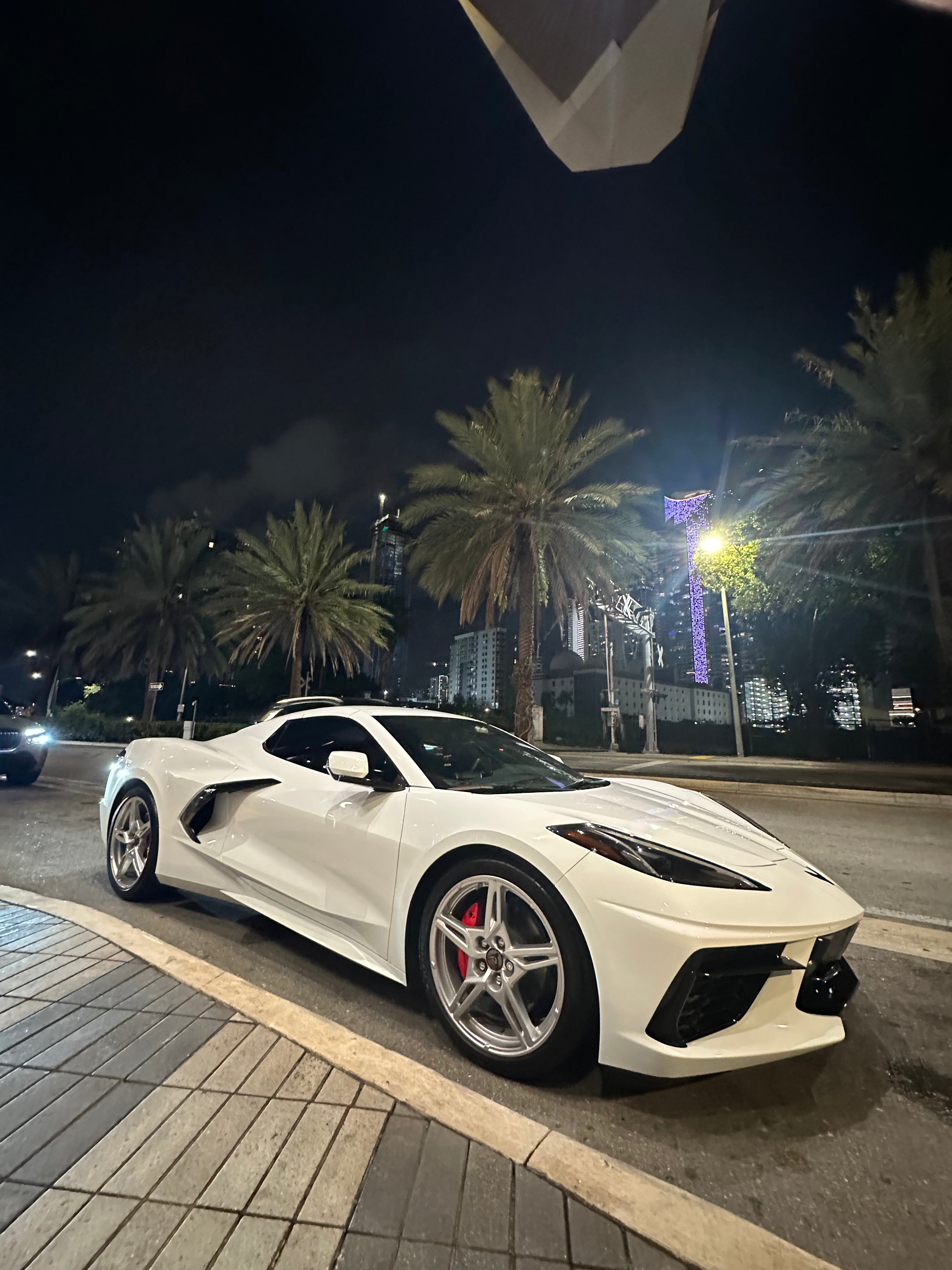 White sports car parked on a street at night with palm trees and city lights in the background