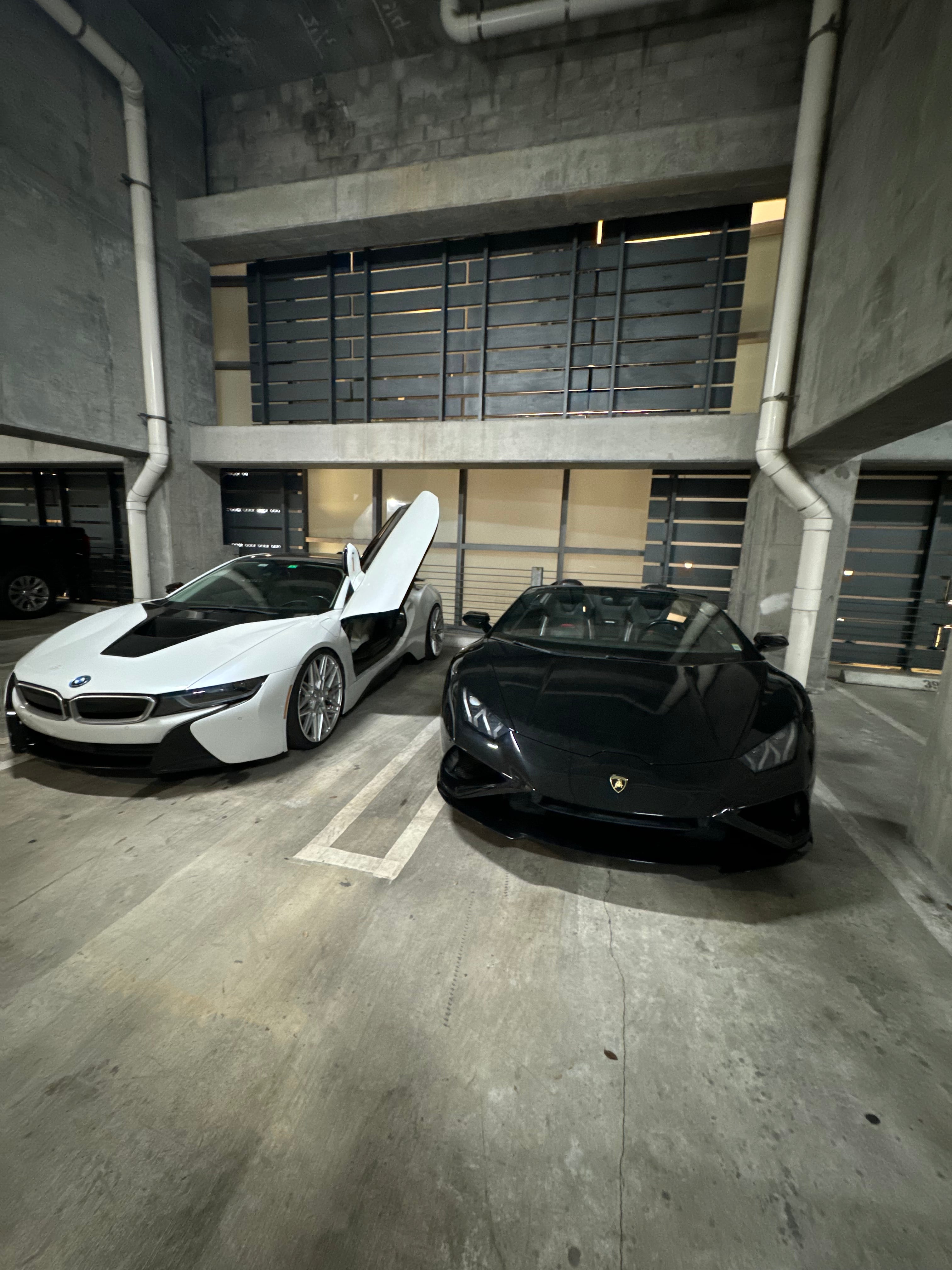 Two luxury cars, a white BMW i8 and a black Lamborghini, parked in a concrete garage.