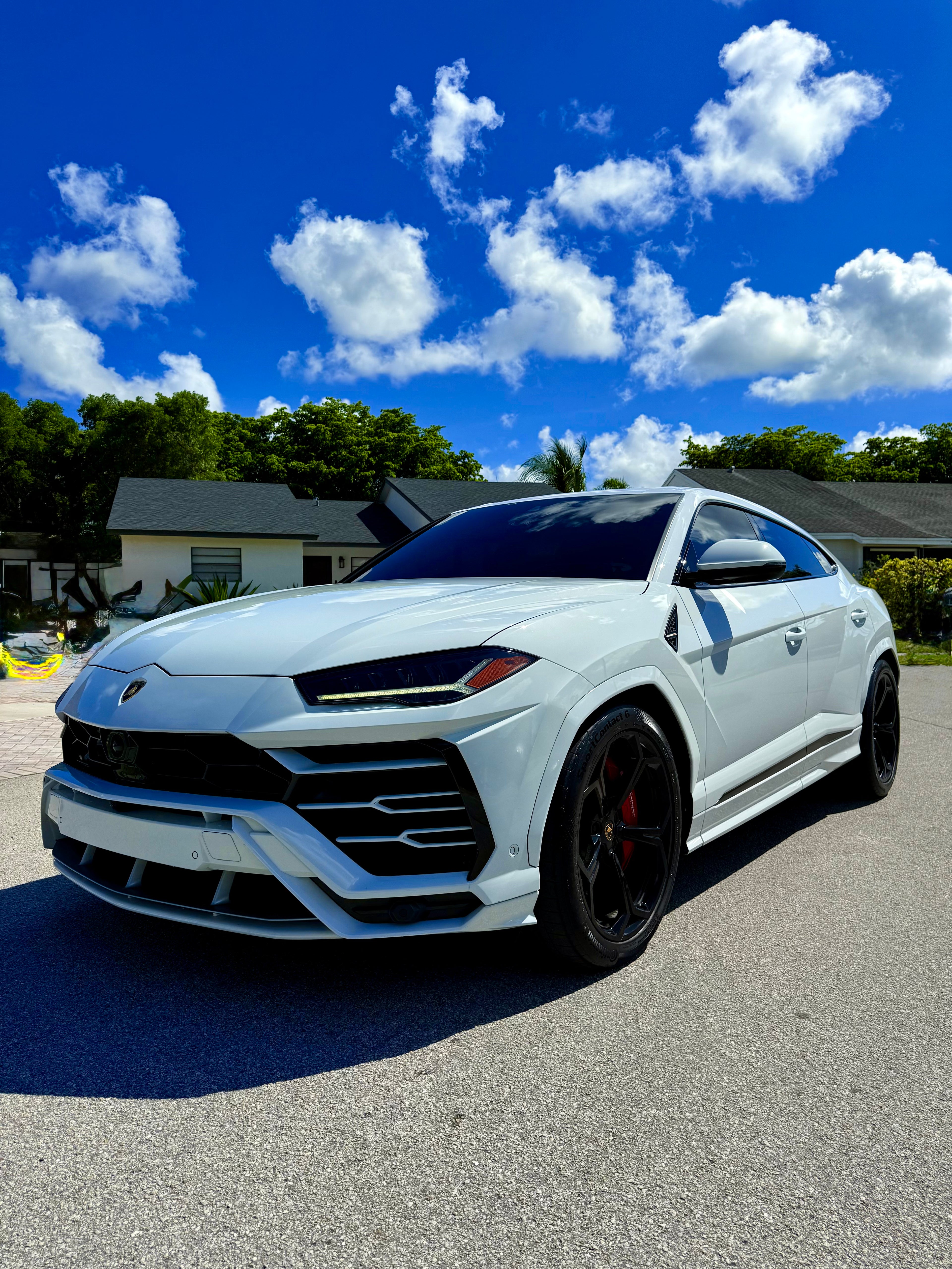 White Lamborghini Urus parked on a street with a blue sky and clouds in the background.