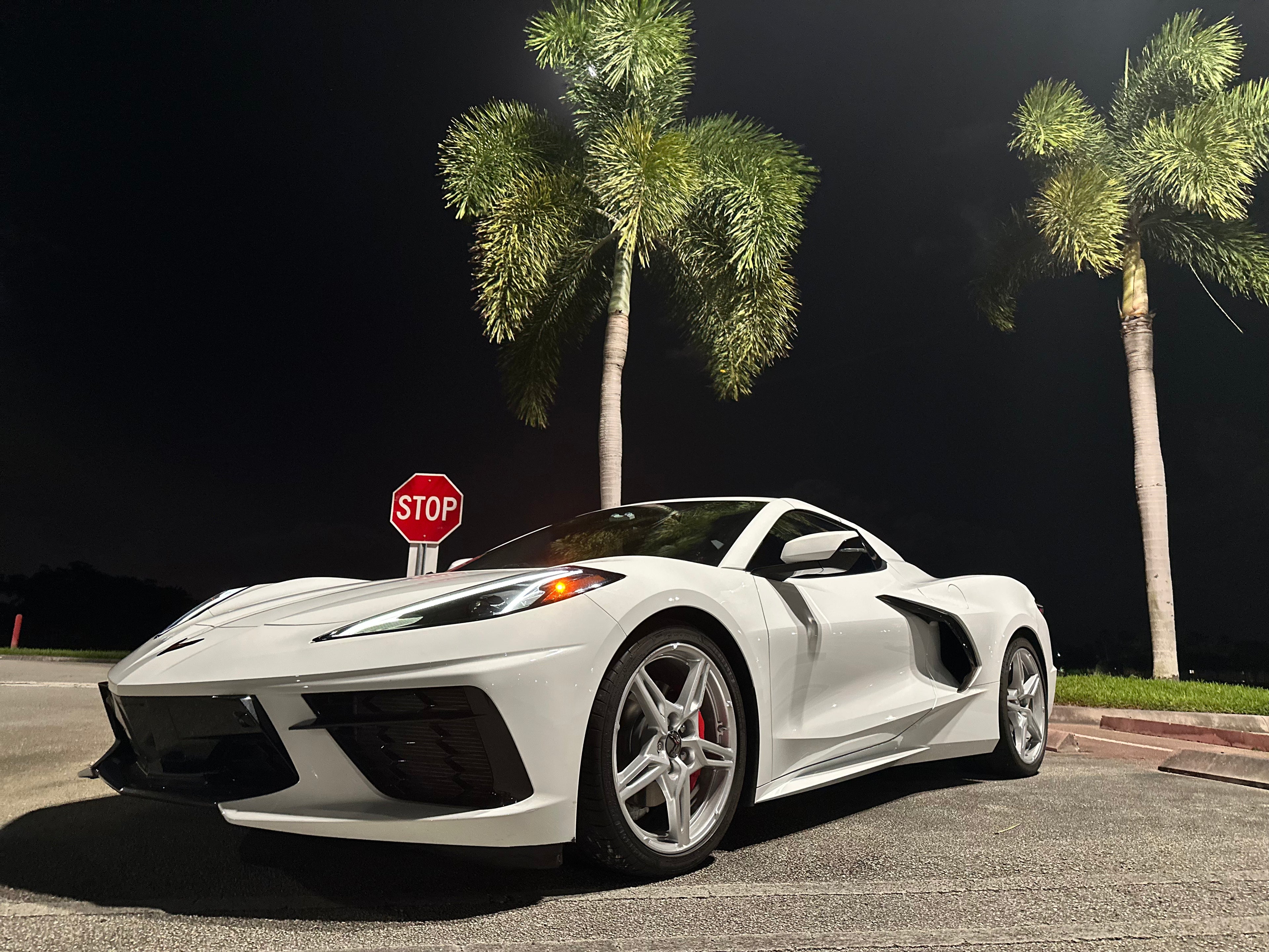 White sports car parked on a road with palm trees and a stop sign in the background.